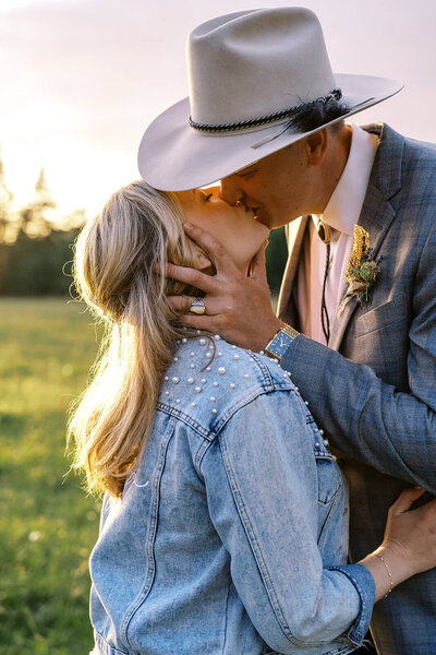 A groom wearing a cowboy hat kisses a bride in a denim jacket.