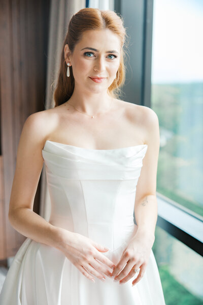 Bride with red hair stands in cat eye basque waisted gown next to hotel window