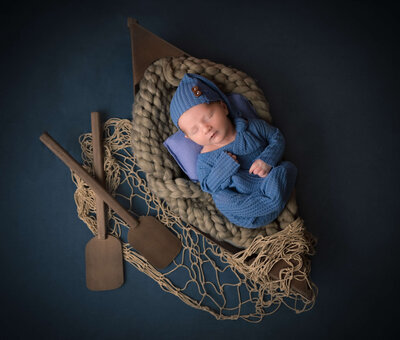 In home newborn photo session by E. Benecki Photography with baby  peacefully sleeping in a fishing boat with oars and fishing net on a dark blue background.