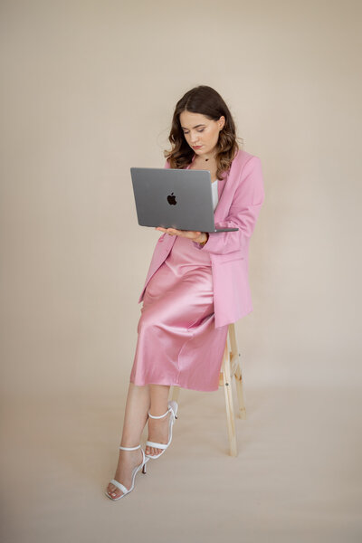 Picture of the Shapely Studio founder dressed in pink, looking. at the laptop while leaning on the chair