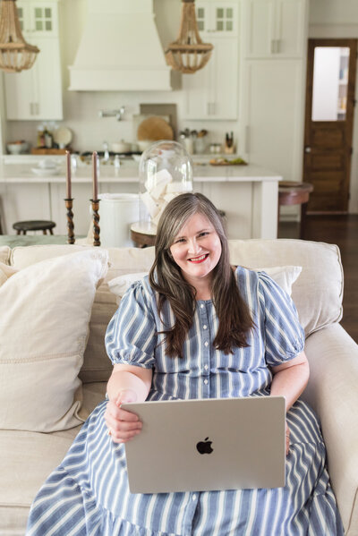 Lauren sits on her family room sofa in white and blue striped dress with her laptop