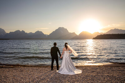 A bride and groom holding hands at the shore of Jackson Lake with the Teton Range in the background, the bride's veil blowing in the wind as the sun sets behind them.