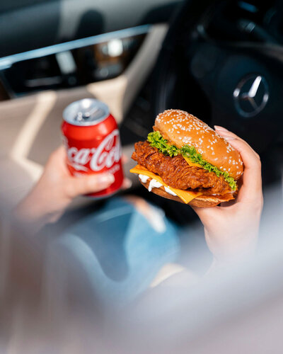 person sitting in their car holding a chicken sandwich and a canned drink