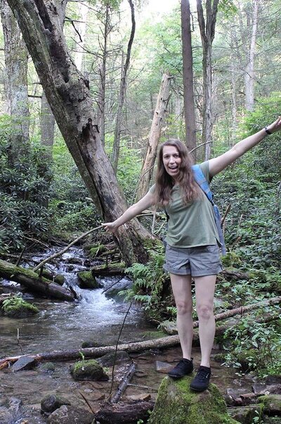 Stacey Xanthe smiling with arms outstretched while standing on a rock in the Roaring Run creek at Shingletown Gap trail near Tussey Mountain, wearing a pale green t-shirt and small backpack.