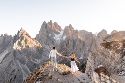 Groom is walking behind the bride. They are at a ski resort in the summer. They are among grass with trees behind. There is a ski lift above them and a mountain in the distance