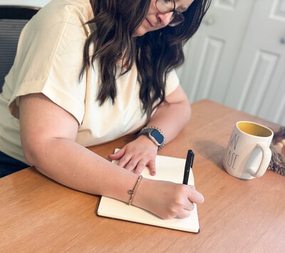 Close-up of Ali's hands writing in a journal next to a coffee mug labeled "sun please"