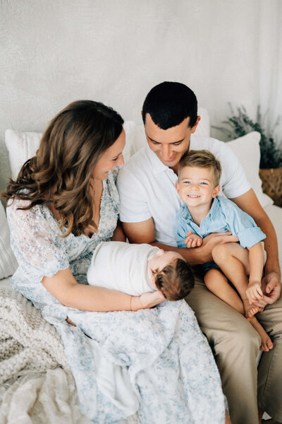 mom holding newborn baby while brother sits in dad's lap and smiles at the Denver photographer