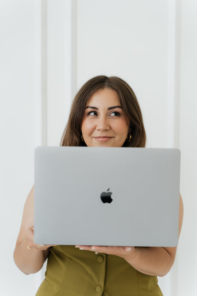 Photographer Darling Darlene smiling behind a silver MacBook, dressed in an olive top, in a minimal white studio.