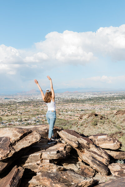 Portrait of a girl on the top of a mountain in Arizona. She is posing with her hands up in the air looking out to the valley. She is wearing blue Levi jeans and a baseball cap.