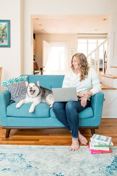 Blonde woman with a laptop sitting on a blue couch with a husky