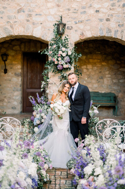 Bride and groom standing in front of stone arches with lavender floral arrangements, elegant Scottsdale wedding photography.