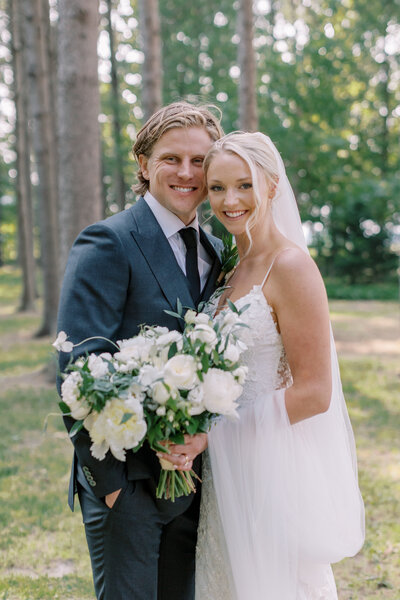 A beautiful bride holding an oversized bridal bouquet with white flowers, standing next to her new husband at wheatfield estate.