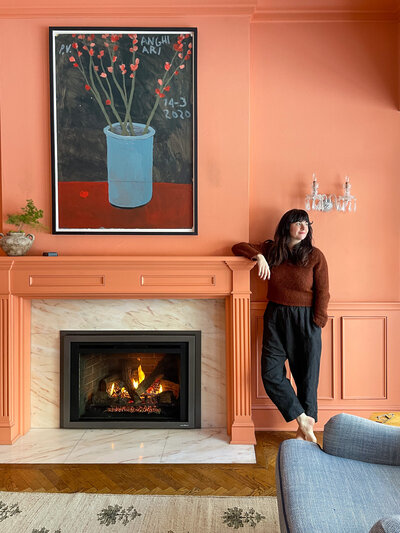 Woman standing by a roaring fireplace in a peach room.
