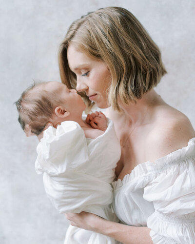 Richmond Newborn Photographer | Film photo of a mom in a white dress holing her newborn son's face gently up to her own face in front of a hand-painted water color backdrop by Jacqueline Aimee Portraits