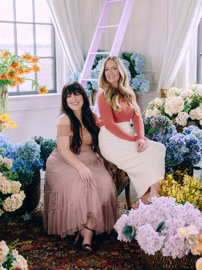 Two women sitting surrounded by buckets of flowers