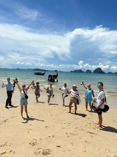A group of people stand on a sandy beach, raising drinks in celebration, with longtail boats and distant islands visible in the background under a bright, cloudy sky.