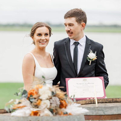 Newly married couple showing their marriage certificate off after their wedding ceremony. Bridal bouquet in foreground with orange and cream florals