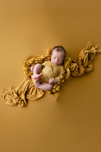 newborn baby boy wearing a yellow onsie laying in a heart shaped bowl on a yellow background for his newborn photography session in Burlington, Ontario.