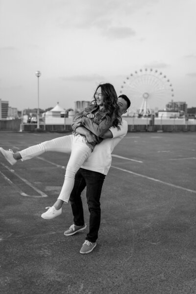 J picking up J from behind and swinging her around, laughing, on the rooftop of a parking garage with the ferris wheel in the background