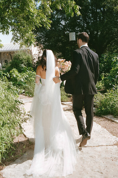Bride and groom walking down a path while groom holds the train of the bride's dress