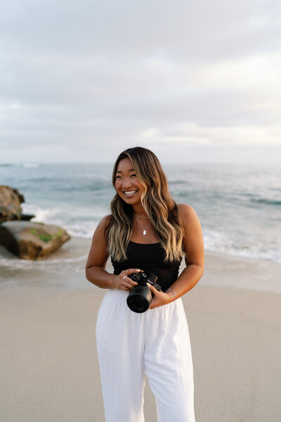 Lauren Ahn, Southern California wedding photographer, smiling and holding a camera while standing on a sandy beach in San Diego, with the ocean waves and rocks in the background, capturing a relaxed and joyful moment.