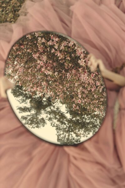 An oval mirror sits atop mauve tulle with the reflection of trees with beautiful pink flowers