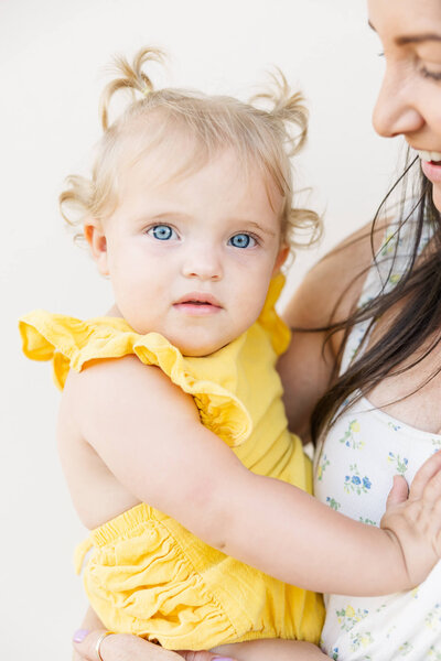A little girl wearing a yellow dress, being held by her mum while she looks directly towards the camera with gorgeous blue eyes.