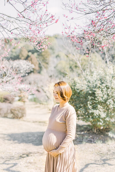 梅の花の下でおなかに手を添えて微笑んでいる妊婦さんのおしゃれで美しいマタニティフォト
