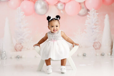 A smiling baby girl sits on a white chair, dressed in an adorable white dress with a pink ribbon. The background features pink and silver balloons, creating a soft and sweet atmosphere perfect for the occasion.