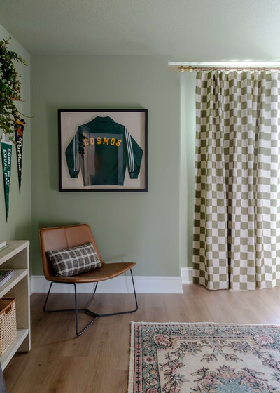 An example of interior design and eclectic home decor, featuring a framed sports jacket on the wall, checkered curtains and a leather chair in the corner. 