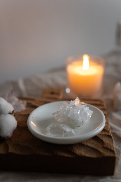 Two pieces of crystal quartz sit in a white ceramic dish on a wooden board atop a table with a lit candle. Learn more about Usui Reiki with The Intuitive Momma.