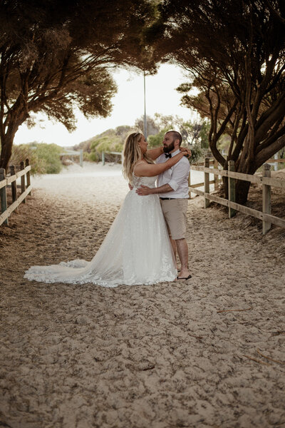 Black & white of wife standing in front of her husband & playfully moving their arms around while holding hands with the beach behind them