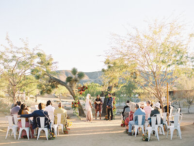 A bride and groom sitting at their reception table looking at eachother and reacting to some speeches. There are wine glasses and food on the table, and more wedding guests behind them. There are market lights above them, and the photo is a black and white film image