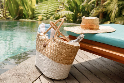 Straw beach bag and sun hat beside a lounge chair by a tropical pool surrounded by lush greenery.