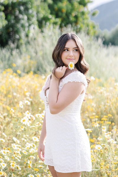 A brown haired young woman wearing a white dress leaning against a wooden gate while looking at the camera
