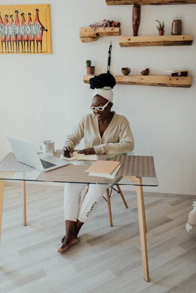 black woman sitting at table writing on notepad with laptop in front of her.