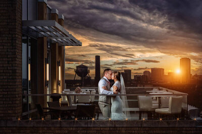 A bride and groom stand on a terrace at sunset, embracing under a partly cloudy sky. The bride's white gown contrasts with the groom's blue suit.
