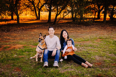 Family Photographer, family of 3 are sitting on grass smiling with their dog