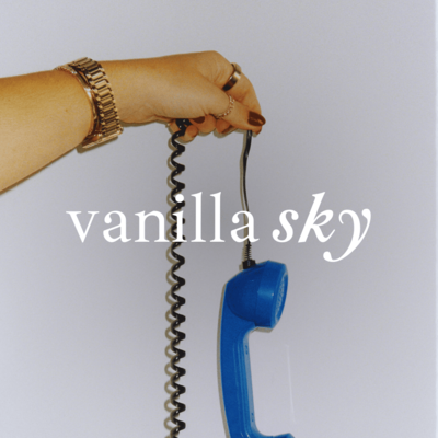 Close-up of a hand wearing a gold watch and rings, holding a blue rotary phone handset with a black coiled cord. The background is a plain wall, and the text 'vanilla sky' is written in a script font across the center.