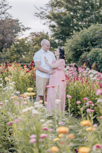 Pregnant woman in pink dress looks up adoringly at her husband in flower garden.