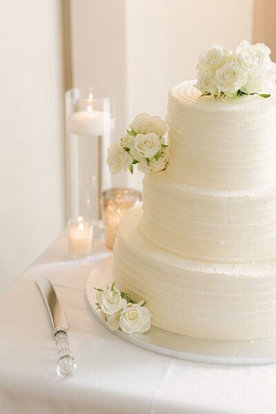 A classic three-tiered white wedding cake adorned with delicate white roses, set against a softly lit table with floating candles and crystal details at Montage Palmetto Bluff, exuding timeless elegance and sophistication.
