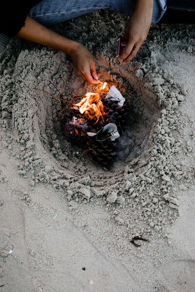 Jemand sitzt am Strand auf dem Boden und macht ein Feuer in einem Sandloch mit Zapfen und Papier: Gemeinschaft und Wärme inmitten der Natur
