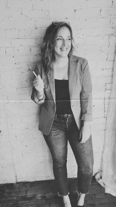 Julie Wenzlick, founder of P&W Designs, smiling in tan blazer and jeans while leaning against white brick wall
