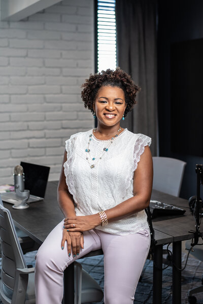 Photo of black woman sitting on desk with podcast equipment in background