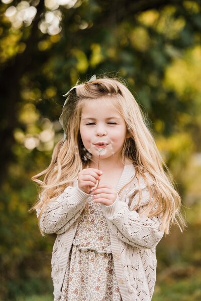 A young girl blowing on a dandelion outdoors, captured by a family photographer.