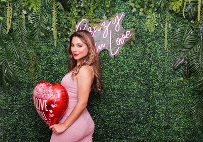 lady holding a red balloon in front a hedge green wall with a neon sign saying crazy in love
