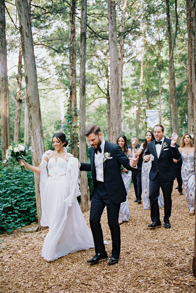 Bride and groom walking hand in hand through a forest aisle at the Caramoor Gardens with their wedding party behind them — a joyful, nature-filled moment captured by Asia Pimentel Photography.