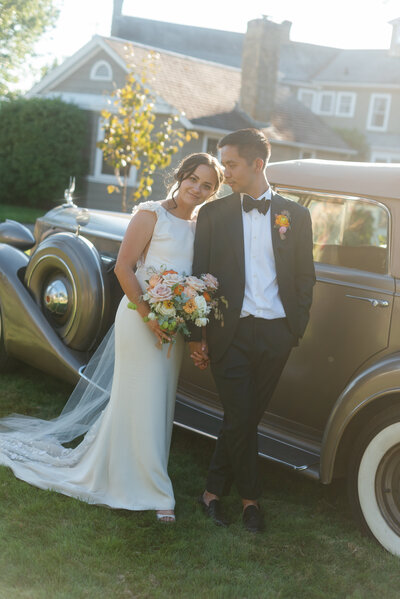 Bride and groom leaning on a vintage car, sharing a quiet moment on their wedding day.