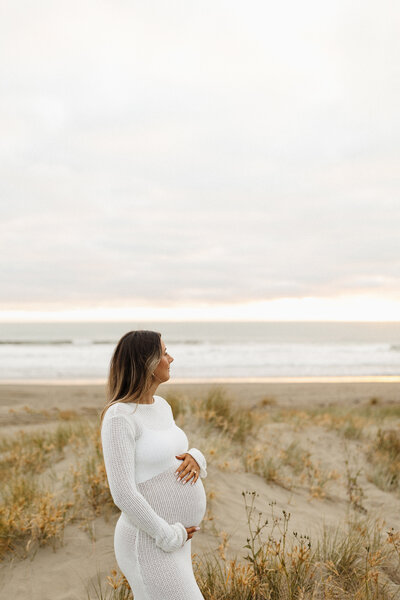 An expectant mother wearing a beautiful white dress from lucy janes client wardrobe for her maternity photoshoot at a christchurch beach