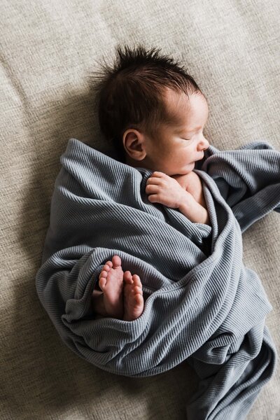 Newborn baby wrapped in a gray blanket, lying on a soft surface during a newborn and family photoshoot.